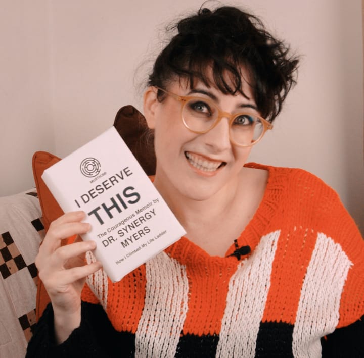A grinning Meljoann holds up a book: "I Deserve This: The Courageous Memoir by Dr. Synergy Myers"